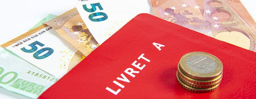 Un livret rouge marqué "Livret A" posé sur des billets de 50 euros et 100 euros avec quelques pièces, illustrant une solution classique d'épargne bancaire en France.