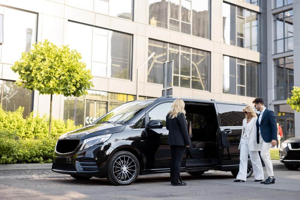 Une chauffeuse professionnelle accueille un couple élégant à bord d'un mini bus avec chauffeur noir devant un immeuble moderne.