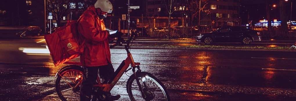 Homme en manteau rouge sur vélo, livraison de nuit en ville sous la pluie