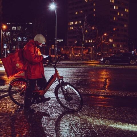 Homme en manteau rouge sur vélo, livraison de nuit en ville sous la pluie