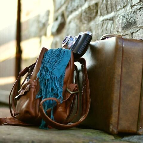 Sac en cuir, écharpe bleue, appareil photo et valise contre mur de pierre